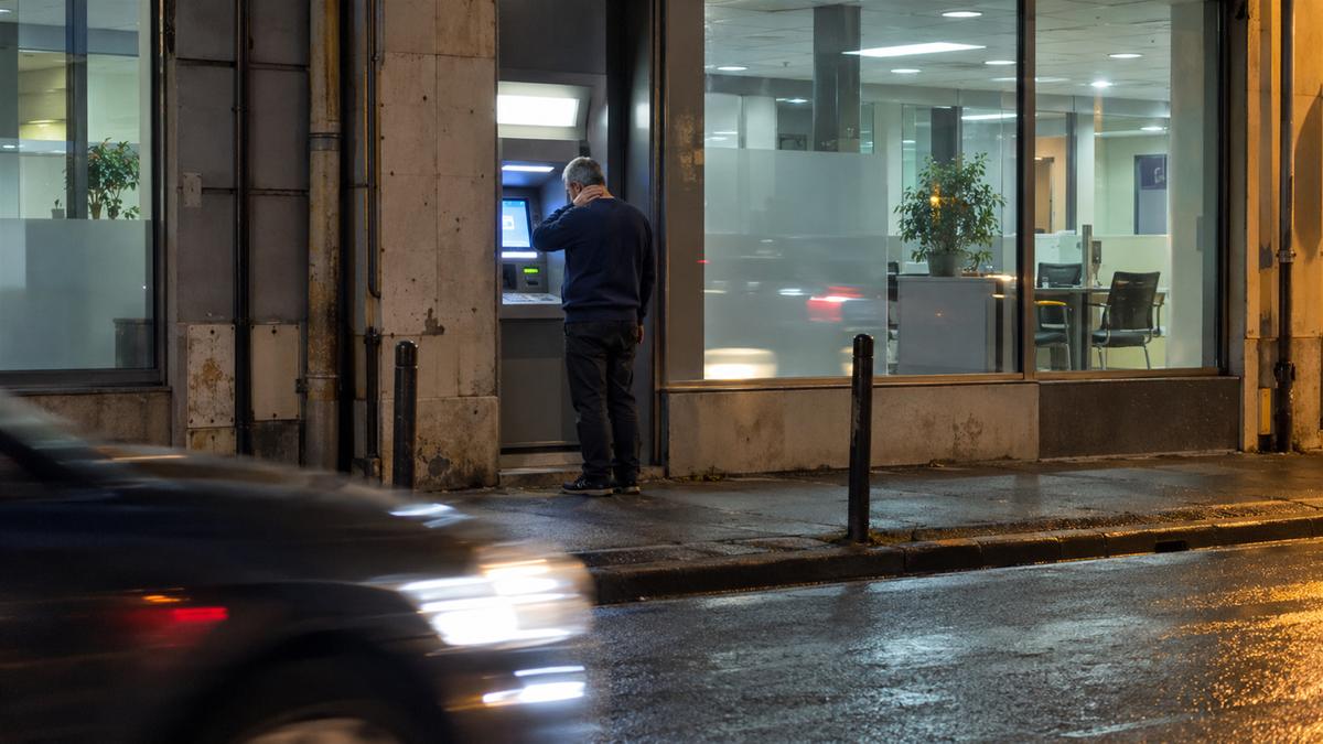 Homme debout devant le distributeur d'une agence bancaire, la nuit, main sur la nuque